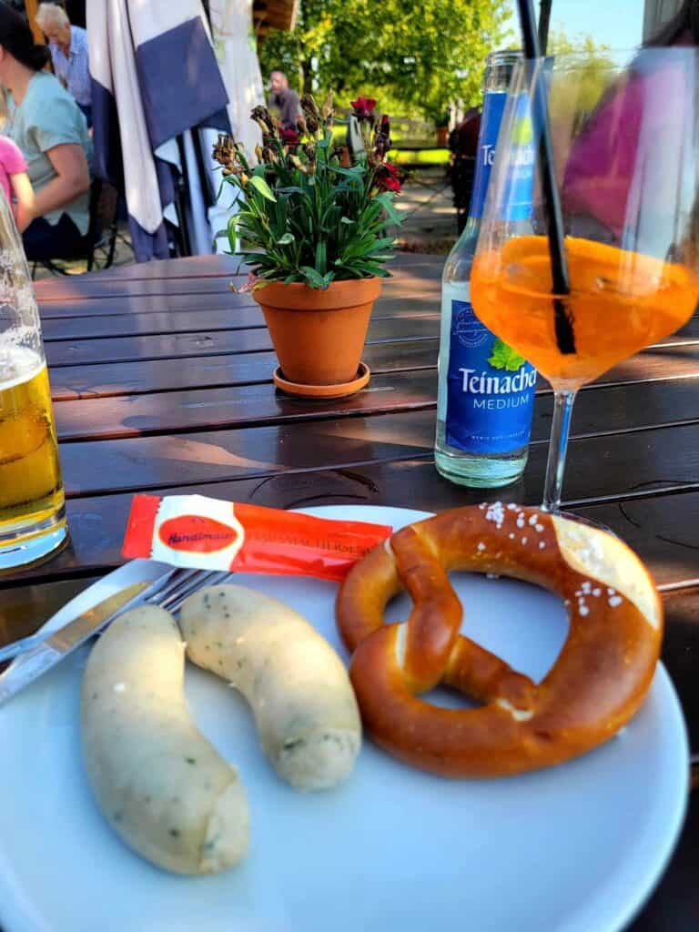 Two weisswursts and a soft pretzel on a plate, an Aperol spritz and a beer on the table