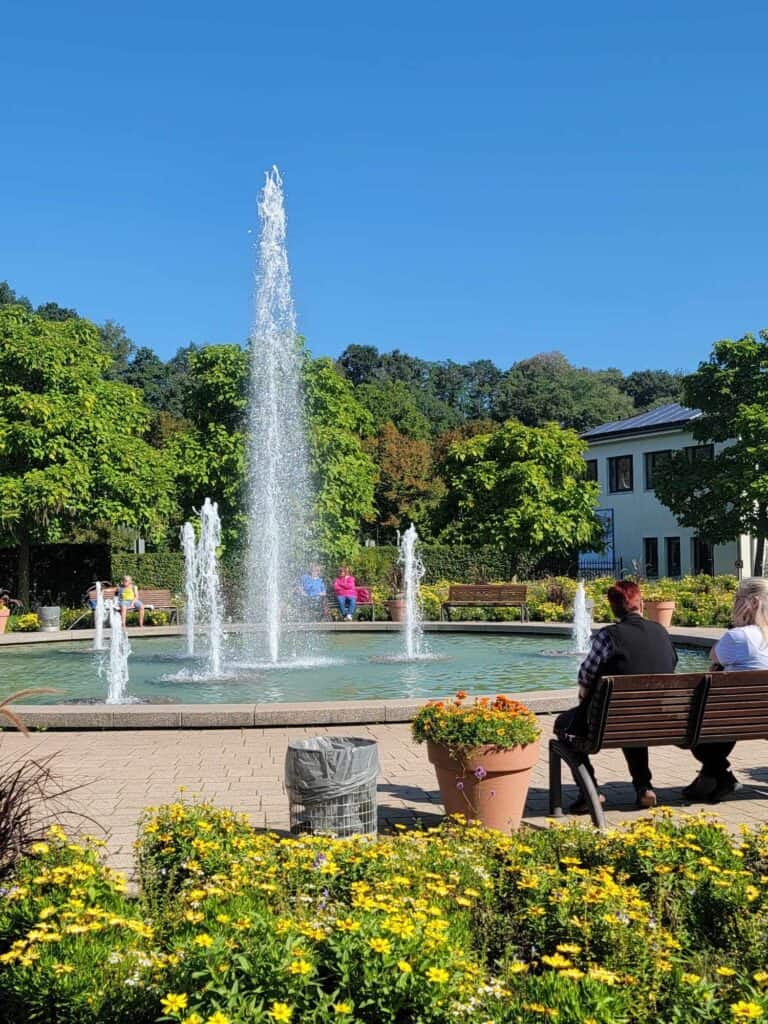 A round fountain with blue water and 8 vertical water spouts. People sitting on benches watching the fountain surrounded by yellow flowers