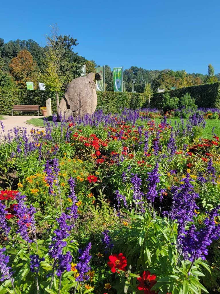 A garden with dark purple and red flowers and a sculpture of a dinosaur hatching from an egg