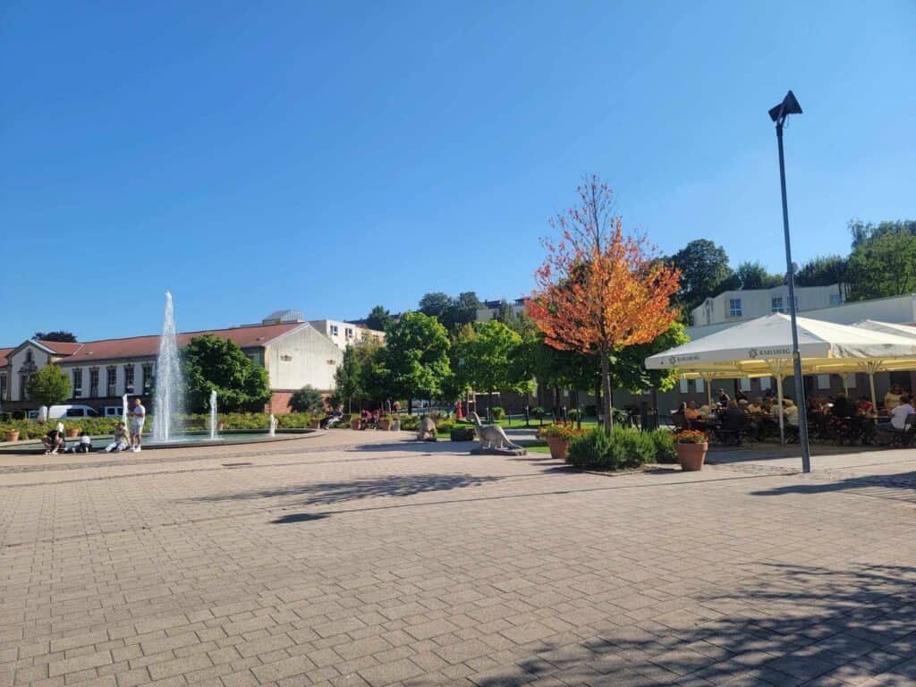 A large plaza with brown pavers, a fountain on the left, trees, and many umbrellas over outdoor tables at a biergarten