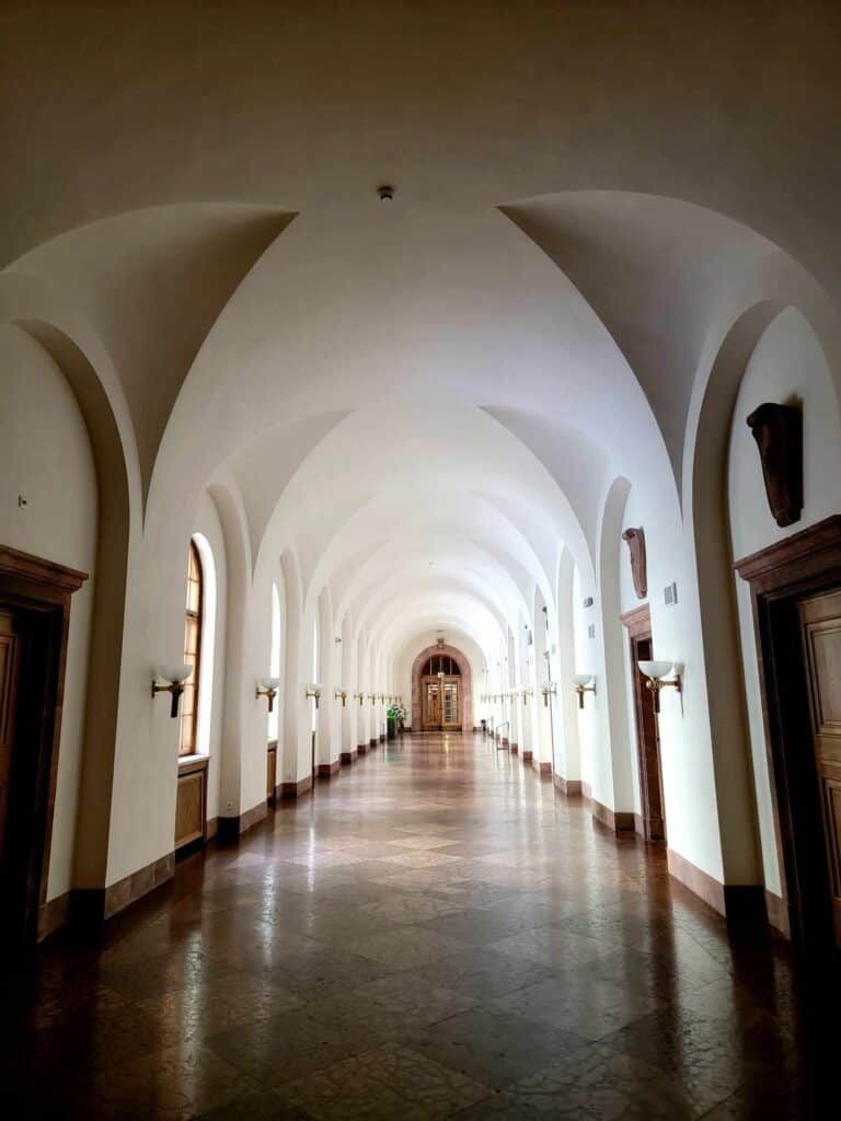 A long arched hallway with a white ceiling, wooden doors and arched windows on each side and a red marble floor