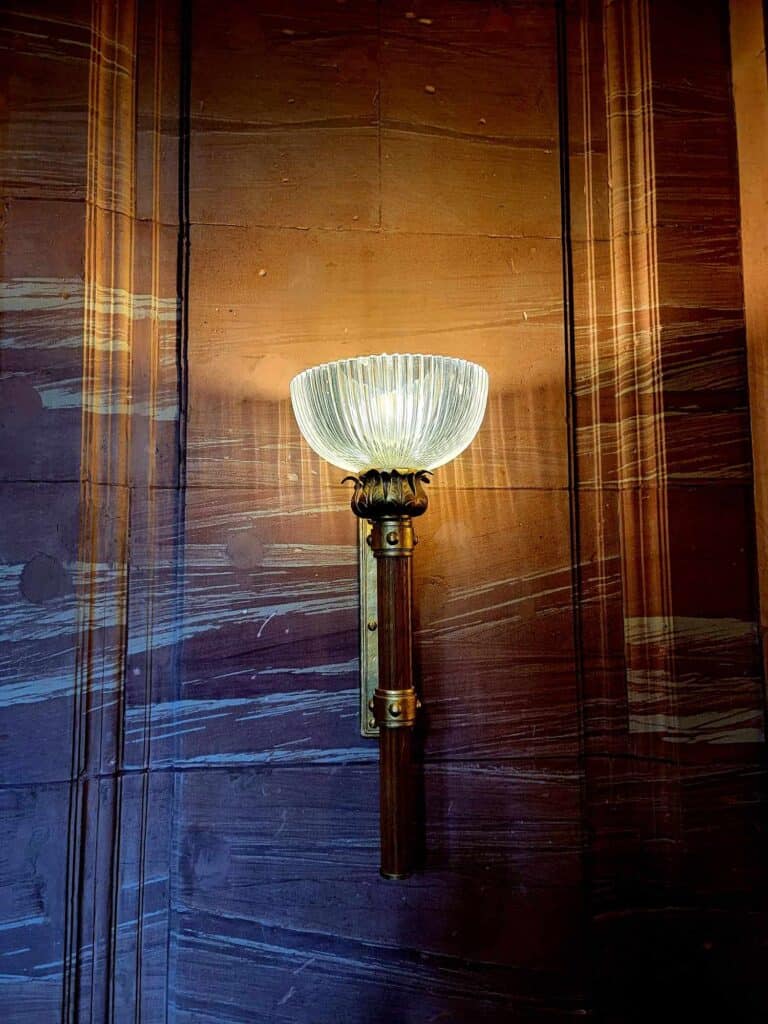 A red and white marble wall with an art deco sconce and filled with large and small bullet holes.