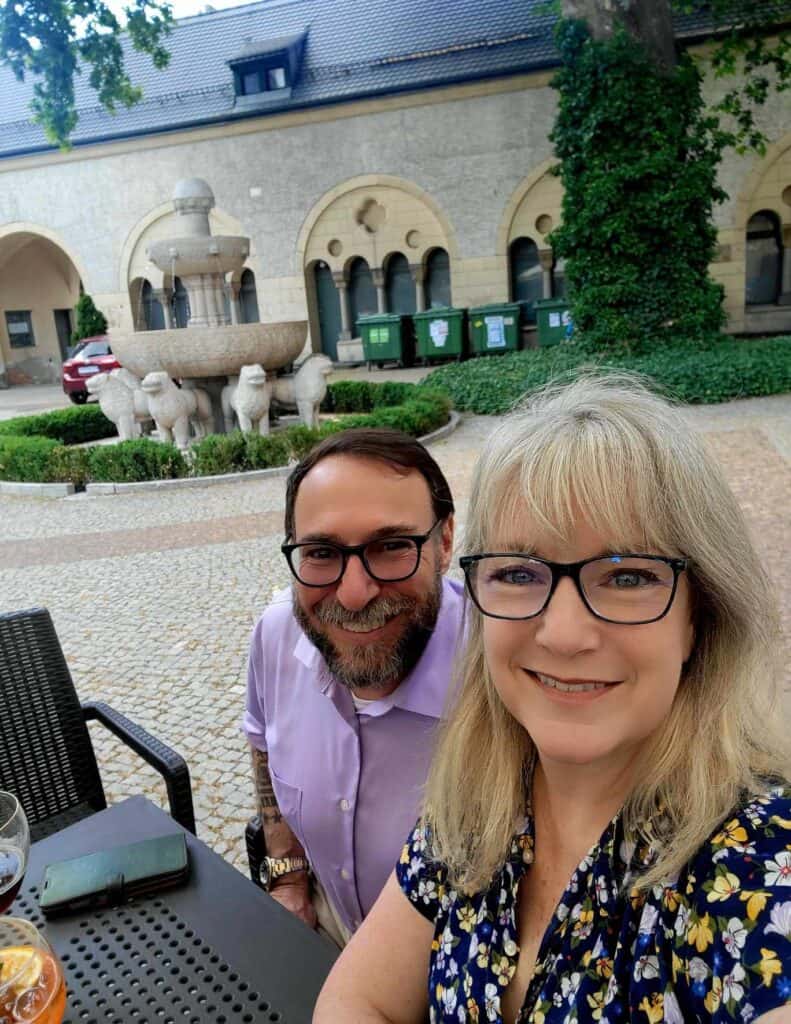 A smiling couple sitting at an outdoor table having drinks with the Lion Fountain behind.