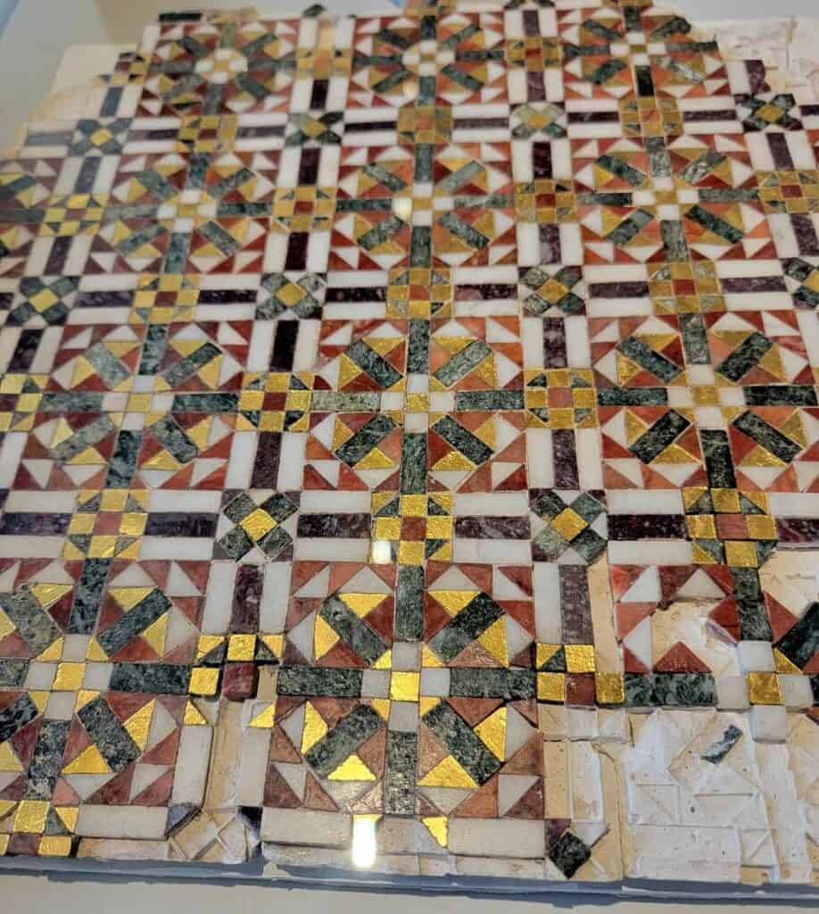 A geometric mosaic fragment in white marble, green and red marble, and gold leaf tiles.