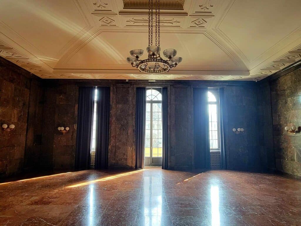 A room with maroon marble floor, brown marble walls, a coffered white ceiling, a chandelier in the art deco style and three tall sets of windows