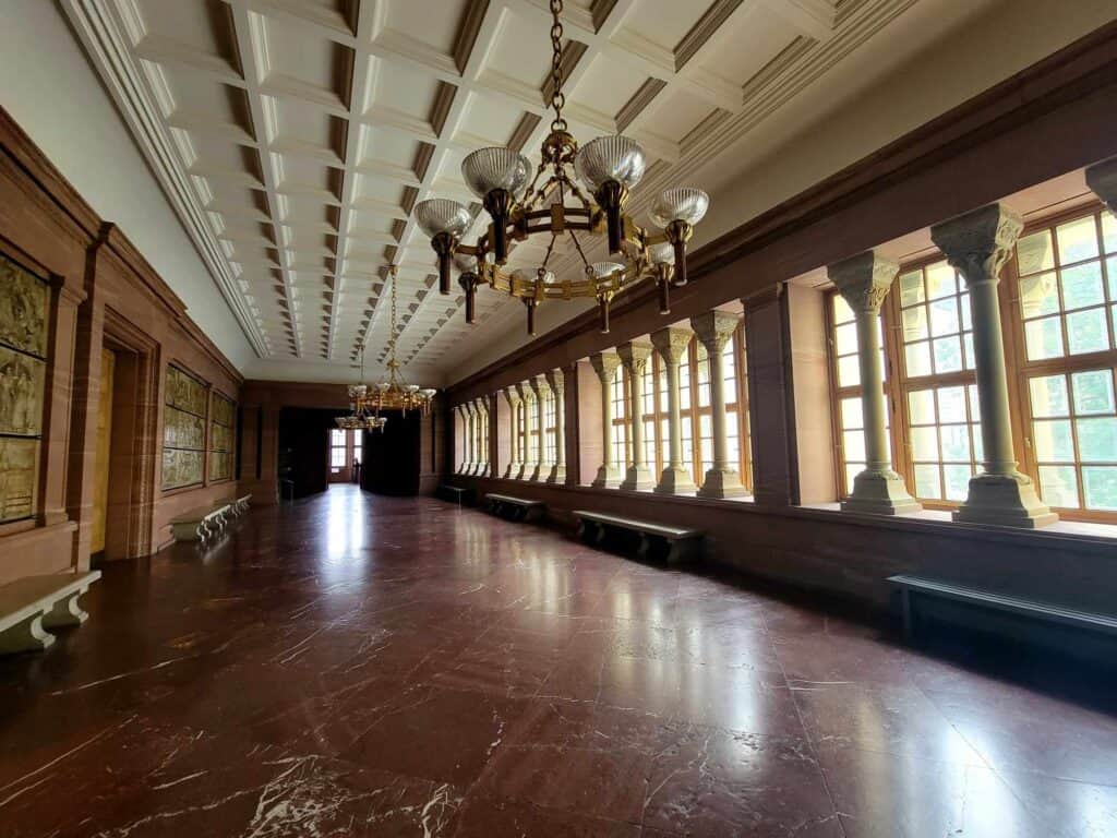 A long corridor with a dark red marble floor, white coffered ceiling, art deco chandelier, and a long row of columns set between windows