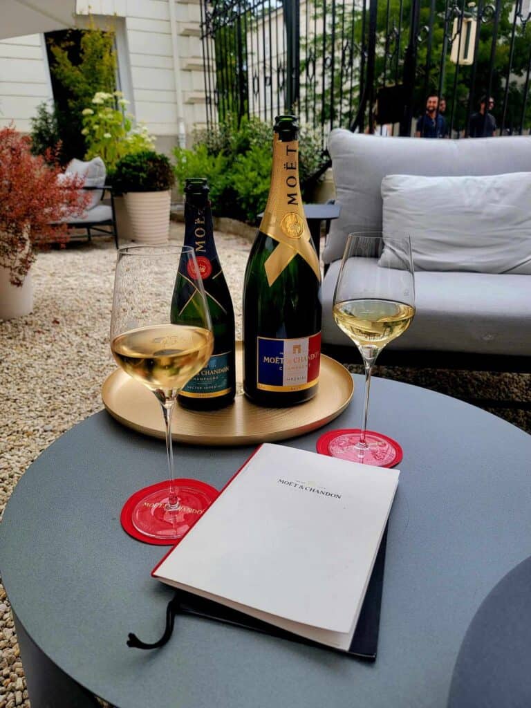 An elegant setting in a garden area with a white chair and pillow and two bottles of Moet and Chandon champagne, two glasses sitting on red coasters, and a menu