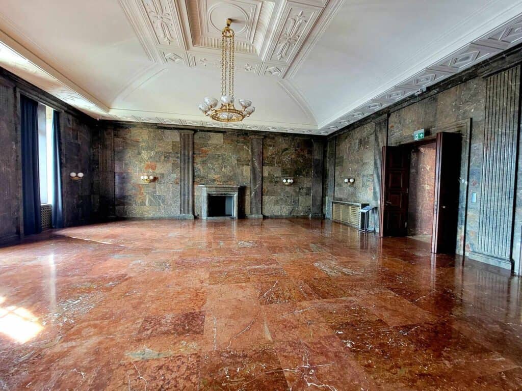 A large room with white coffered ceilings and a mid-century chandelier. The walls are brown marble tile and the floors are red marble. There is no furniture, just a fireplace. This was an office designated for Adolph Hitler, who never made it to the site.