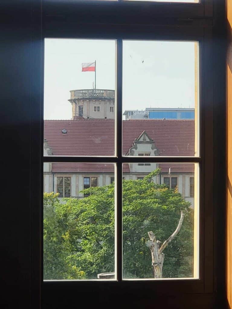 A window with four small glass panes looking out over trees, and a red roof, showing a round tower flying the Polish flag