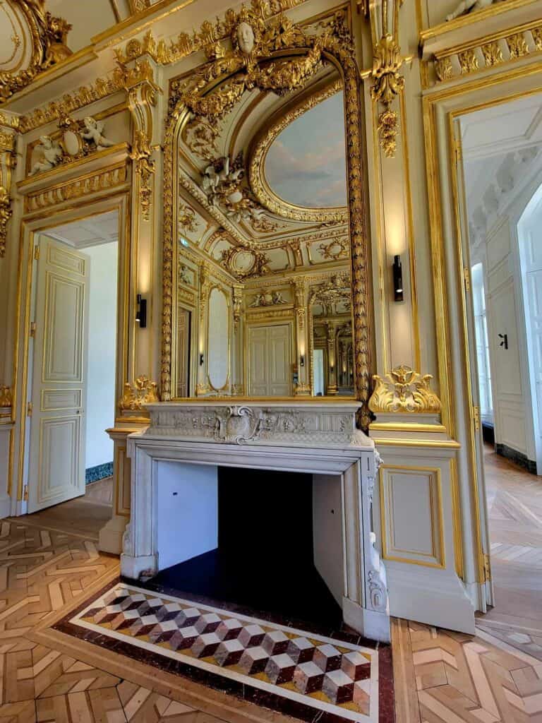 Incredibly ornate gold and white room with baroque detail everywhere and mirrors. A white marble fireplace and a marble geometric floor