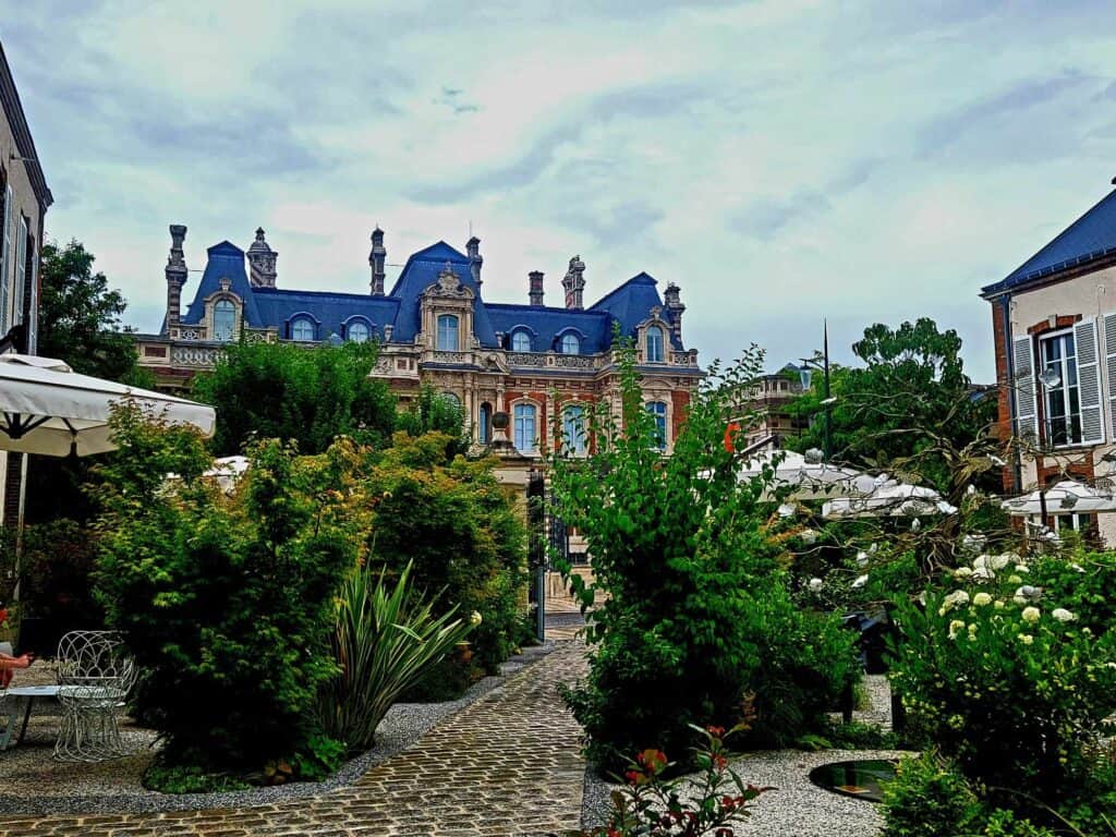 A stone path with luxurious greenery on either side and a french mansion on the other side