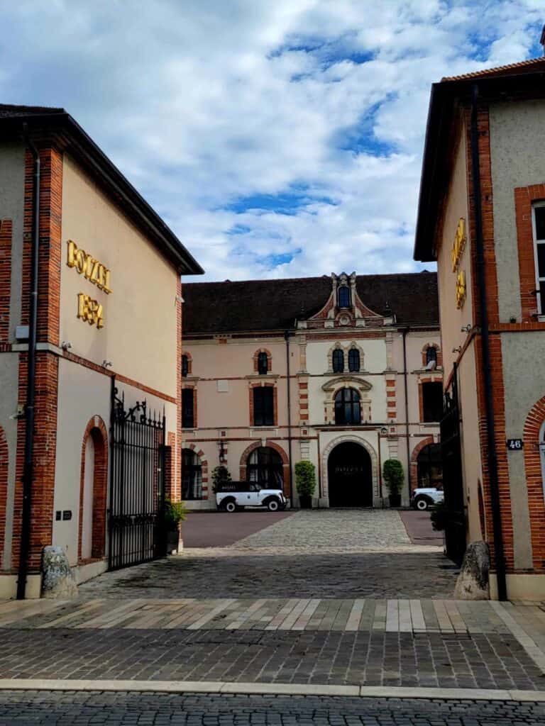 A mansion with two white cars in front, looking through opened iron gates