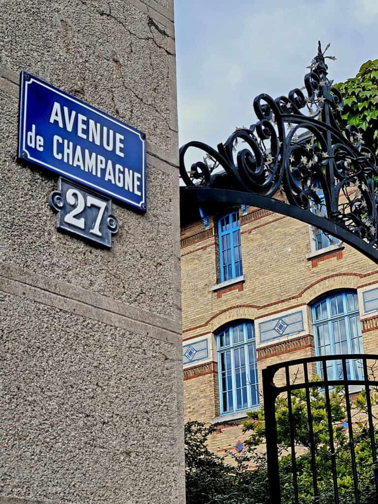 A stone wall with a blue metal street sign that says "Avenue de Champagne" and a wrought iron scrolled gate