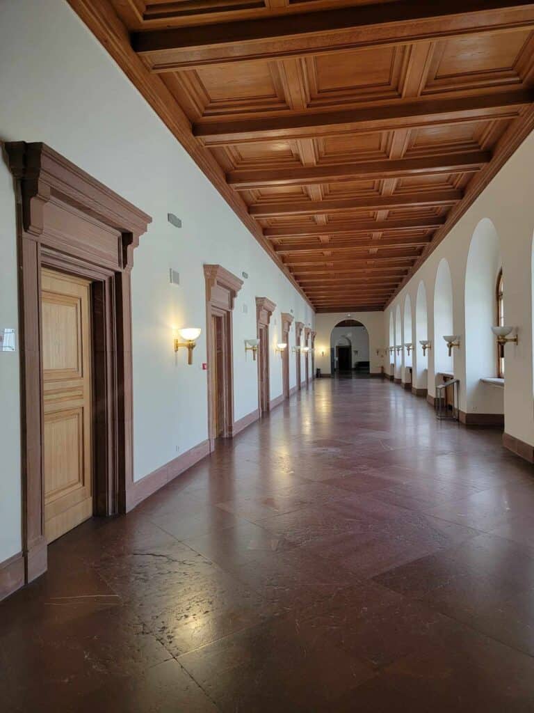 A long corridor with wooden doors on the left, and frosted arched windows on the right, with a coffered wooden ceiling