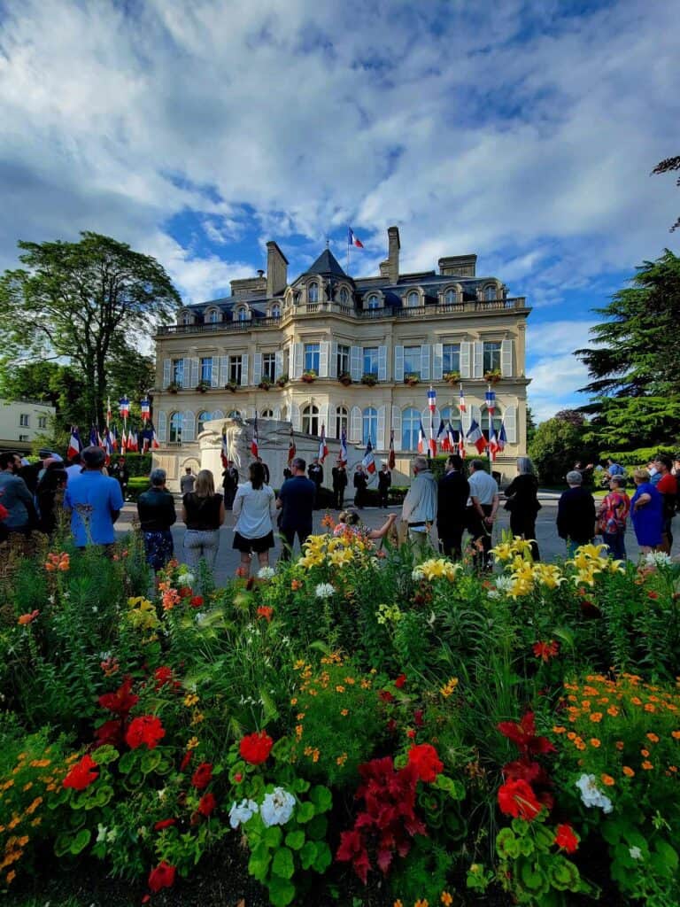 A white 19th century building with the french flag flying. colorful flowers, and people watching a ceremony