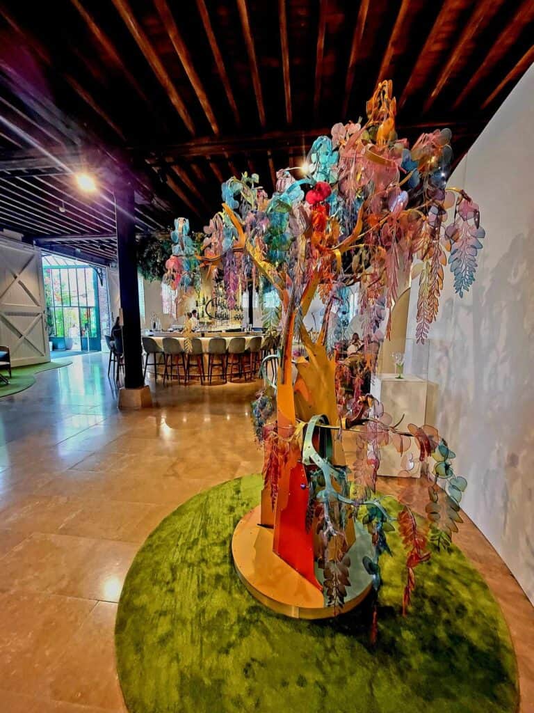 The tasting room of Perrier-Jouët Champagne. A beautiful multicolored glass tree inside a room with a wood beam ceiling, travertine floors and a curved tasting bar surrounded by tall chairs