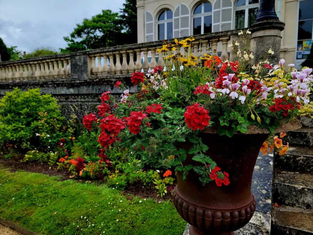 An ornate stone railing and an urn filled with red geraniums and other colorful flowers