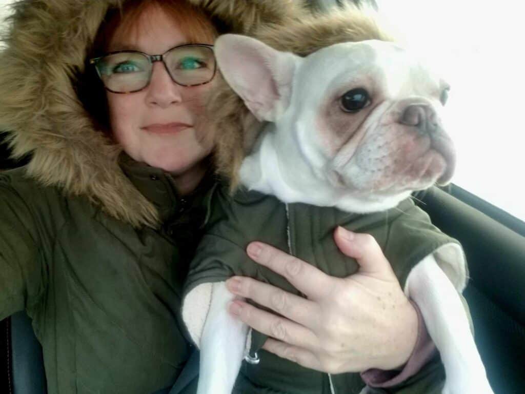 A woman holds a frenchie while they wear matching olive green parkas with ruffed hoods