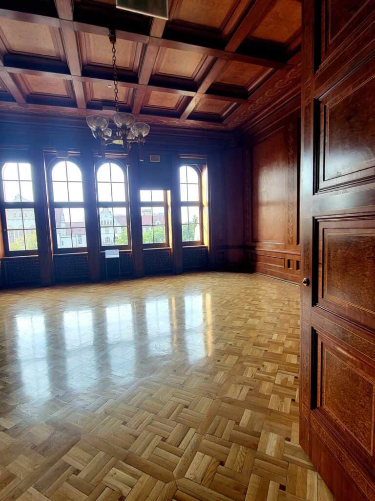 A room with a coffered wooden ceiling, a row of arched windows, a heavy wooden door, wooden paneling, and a parquet floor