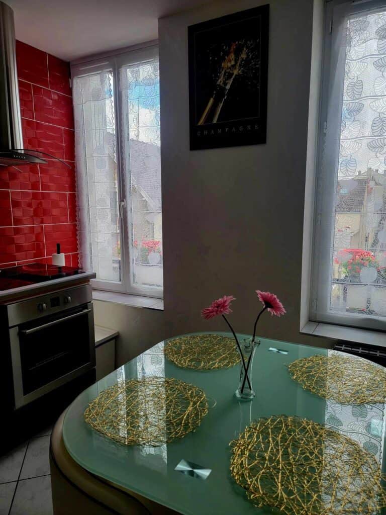 A kitchen with red tile and a turquoise table with four gold placemats and a small vase with 2 gerber daisies. Lace curtains on the windows