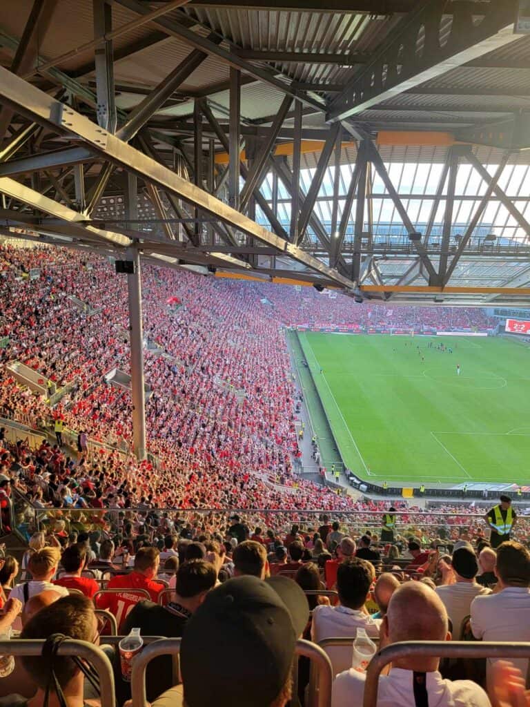 An enormous. stadium full of people wearing red and a green soccer pitch with huge skylight windows overhead