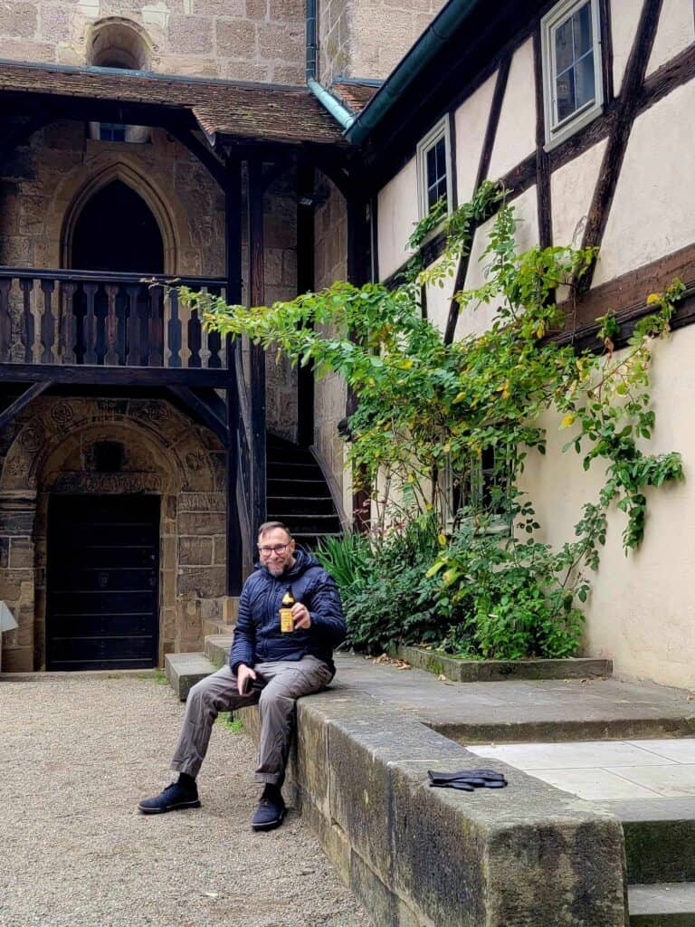 A man sitting in front of a medieval building smiling and holding a bottle of beer