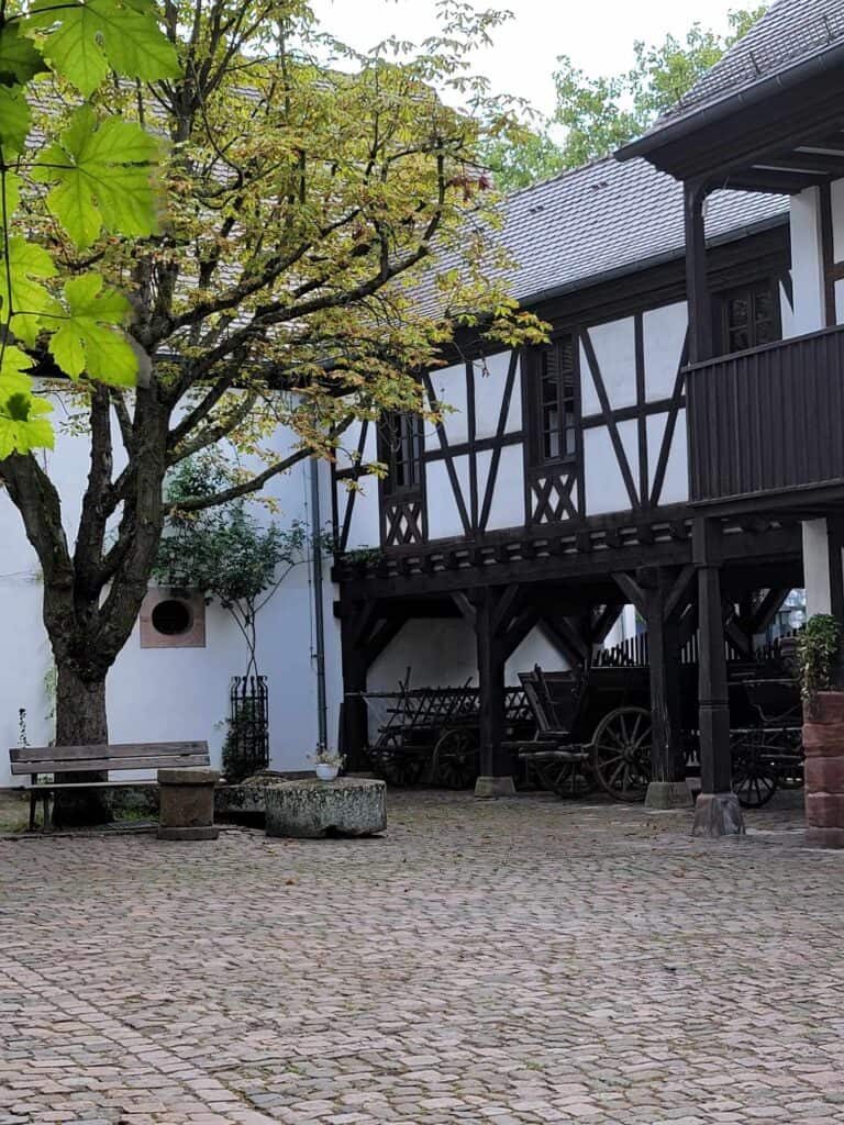 The cobblestonecourtyard of a half timbered house with a tree and a stone basin next to a bench