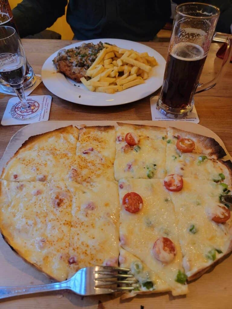 A huge oval-shaped flammkuchen with tomatoes on one side and covered with cheese, two glasses of dark beer and another plate of schnitzel and fries