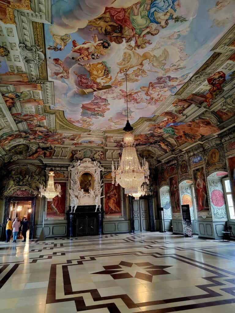 The Kaisersaal, a stunning Baroque room with elaborately frescoed ceiling, a fireplace with a rococo white marble mantle, crystal chandeliers, and a marble floor with star pattern