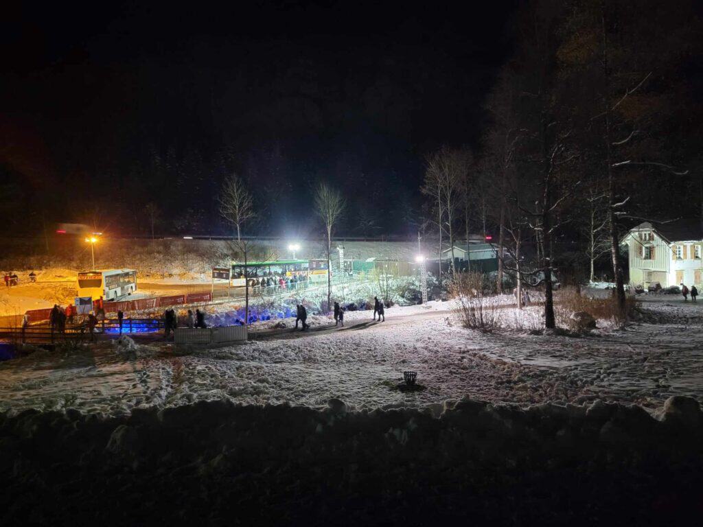 A snowy field with a white building to the right and a parking lot with buses on the left. People are walking to the buses.