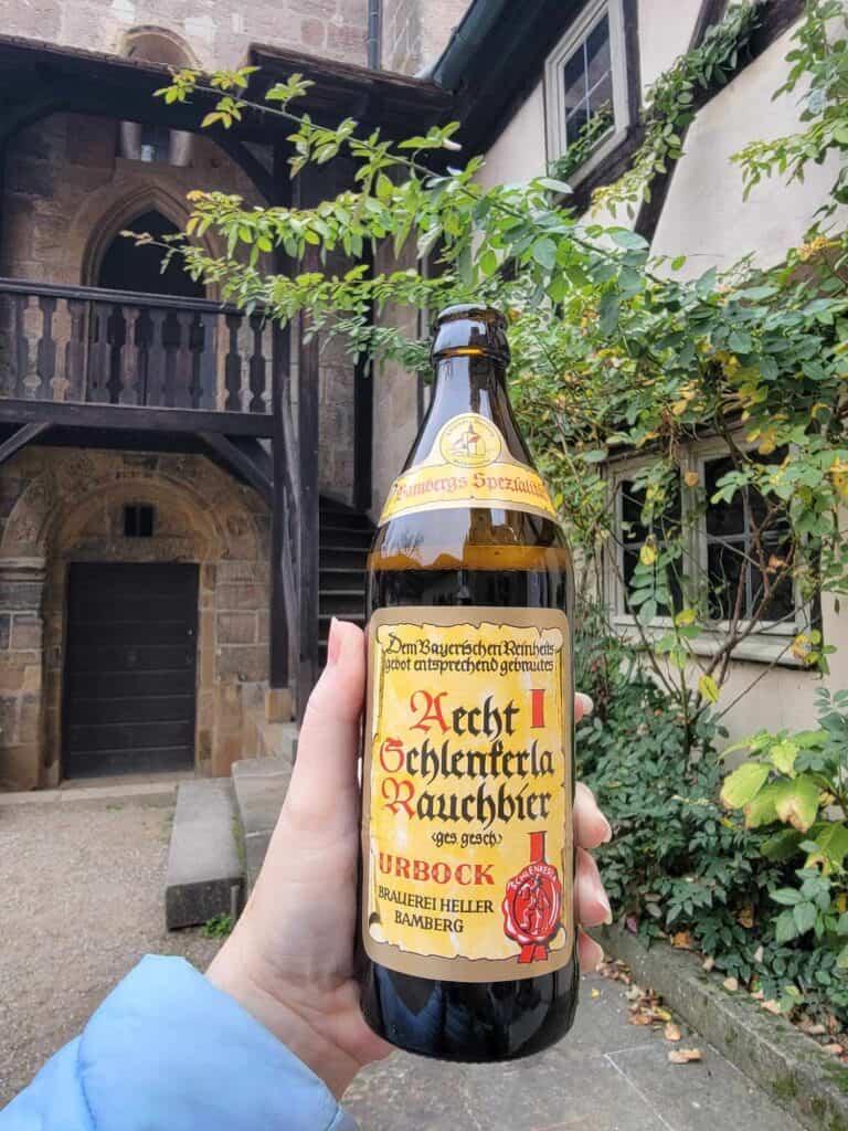 A woman's hand holding a large bottle of Scherlenka Rauchbier with a yellow label with gothic calligraphy. red and black lettering, and a red seal, with medieval building behind