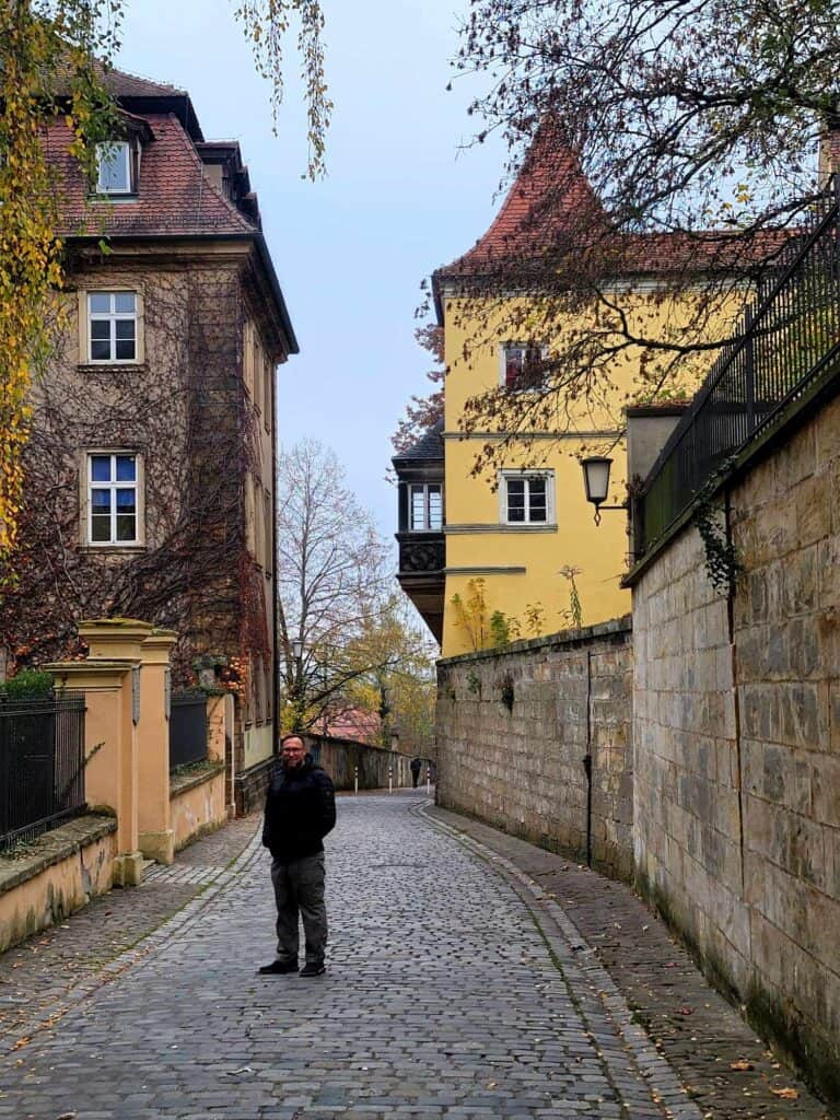 A man on a cobblestone road with a wall on one side and colorful ivy covered buildings