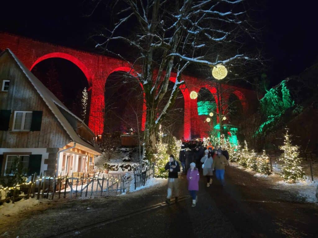 A tall arched stone viaduct illuminated in red with snowy trees and a lane lined with Christmas trees next to a wooden chalet with green shutters. People in heavy coats and hats walk down the lane towards the camera