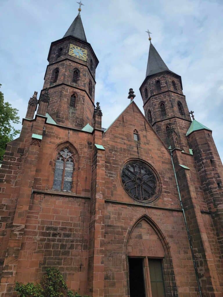 The facade of a red sandstone church with a rose window over a gothic arched doorway and two octagonal towers, one with a clock