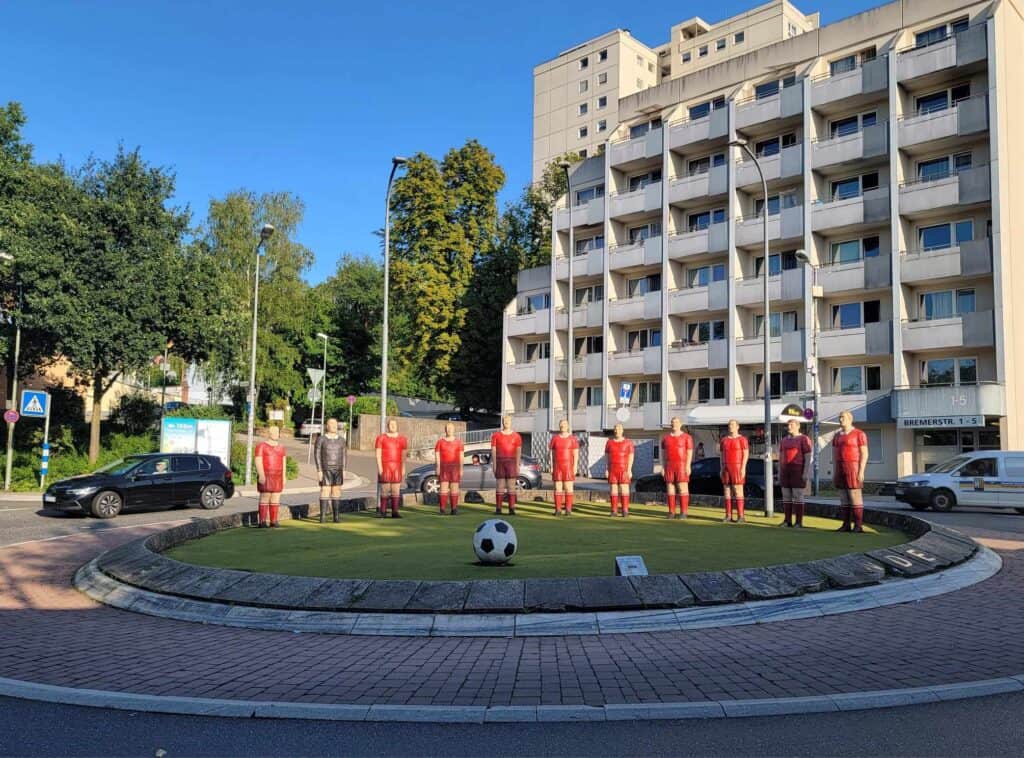 11 life-sized soccer players in red uniforms look at a soccer ball in a traffic circle