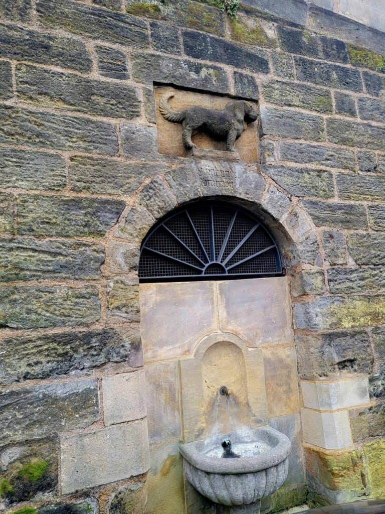 A small fountain in a wall with a carved relief of a small dog overhead