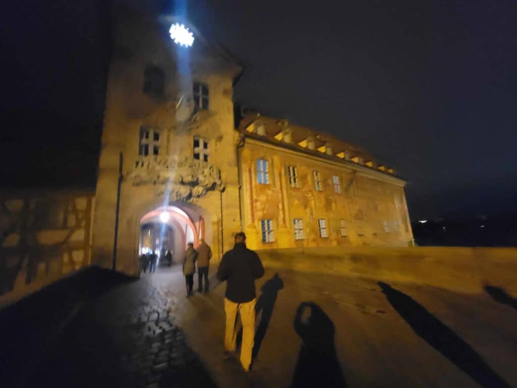 A man at night walking across a stone bridge heading to an arched building