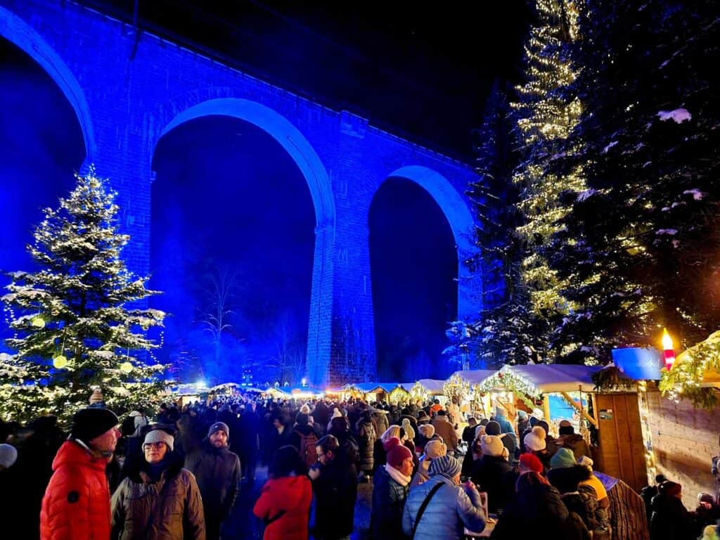 A tall arched stone viaduct illuminated in blue with a long row of market stalls, and two large evergreens with snow and white lights. Hundreds of people in coats and hats browse the stalls