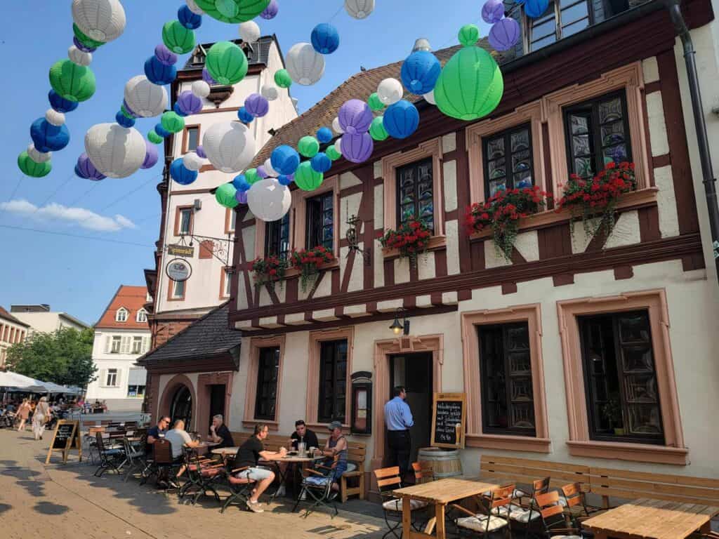 A half timbered building with outdoor tables, and strings of Japanese lanterns over the street in white, blue, green, and lavender