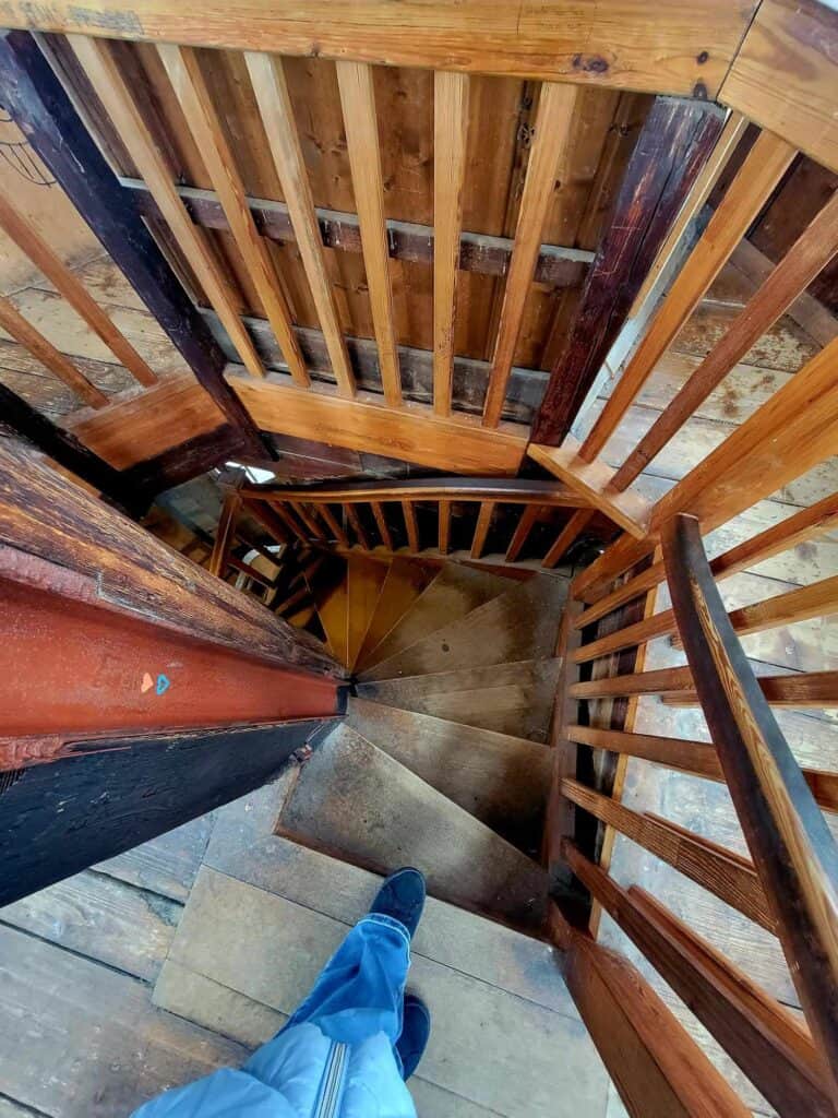 A woman's legs preparing to walk down a very small twisted wooden staircase in a tower