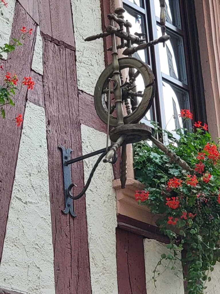 A half timbered building with a small wooden Spinning wheel affixed to the outside and a flower box with red geraniums