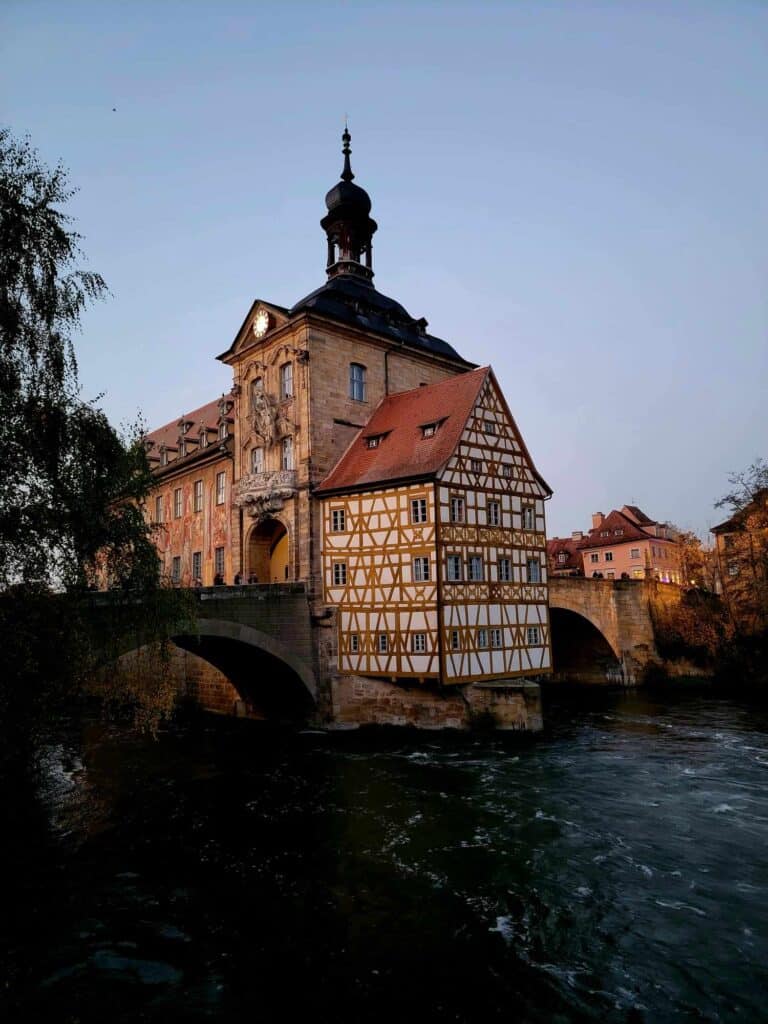 A large building in the middle of a river with an arched stone bridge on either side and a projected room with timber frame.