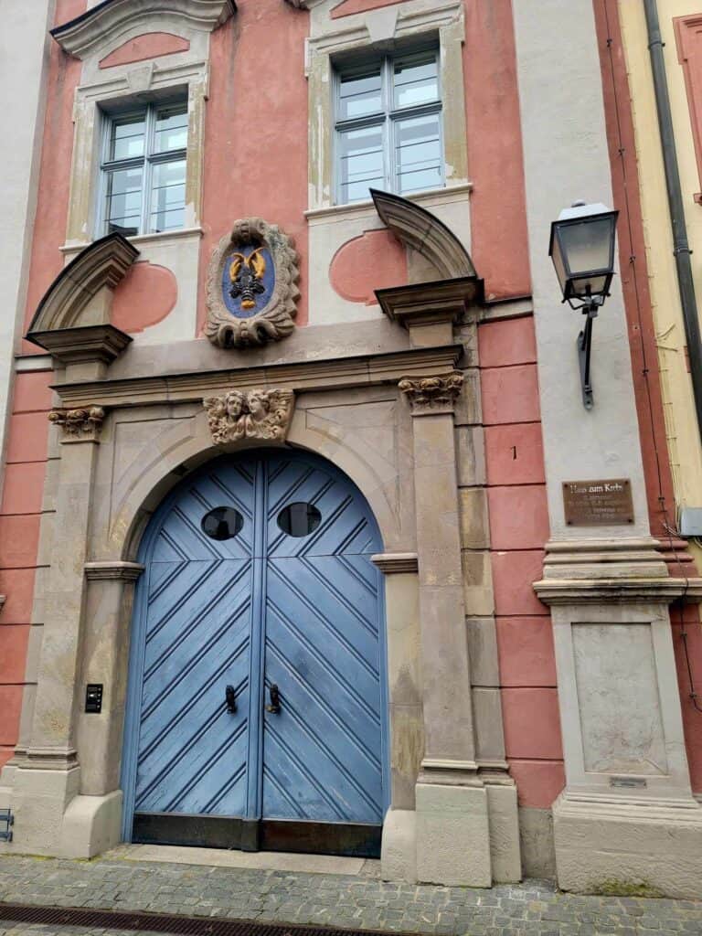 A pink sandstone building with a large blue arched door with oval windows and a carved relief of a lobster over the door