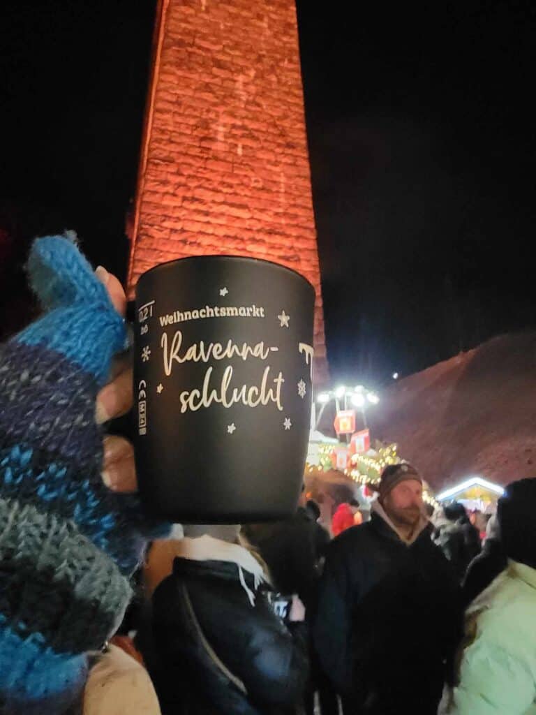 A woman's hand in a fingerless mitten holds a black mug that says Weihnactsmarkt Ravennaschlucht