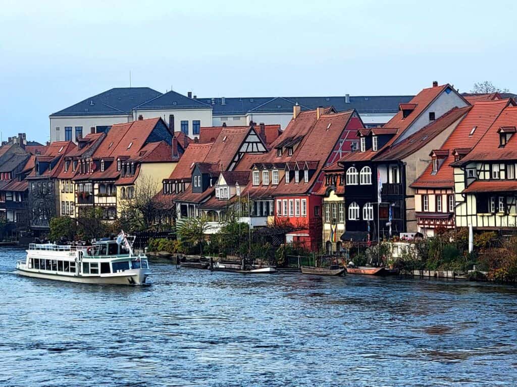 A row of beautiful colorful and half timbered buildings located directlyl on the river with small dinghies floating and a larger tour boat passing