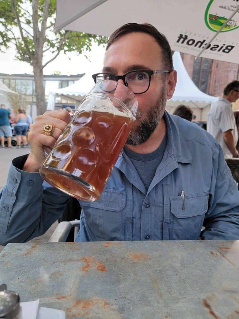 A man sits in the square at a marble table under an umbrella drinking a huge glass of amber beer