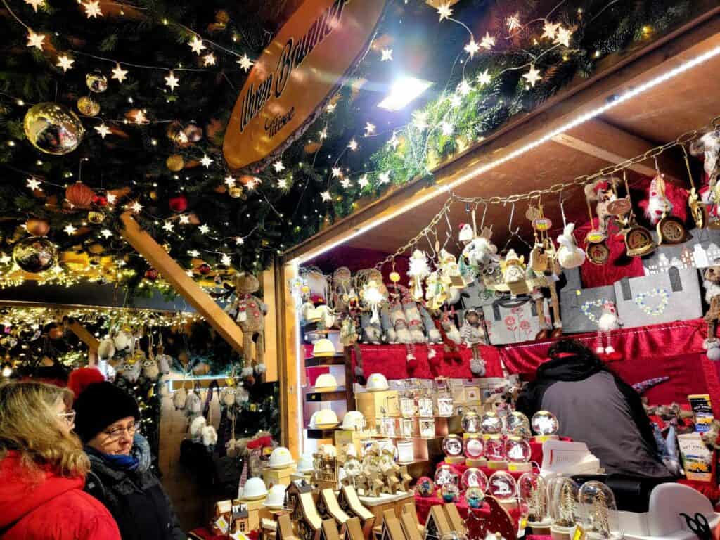 A Christmas market stall full of snow globes and illuminated decorations, with lots of little white lights and greenery overhead