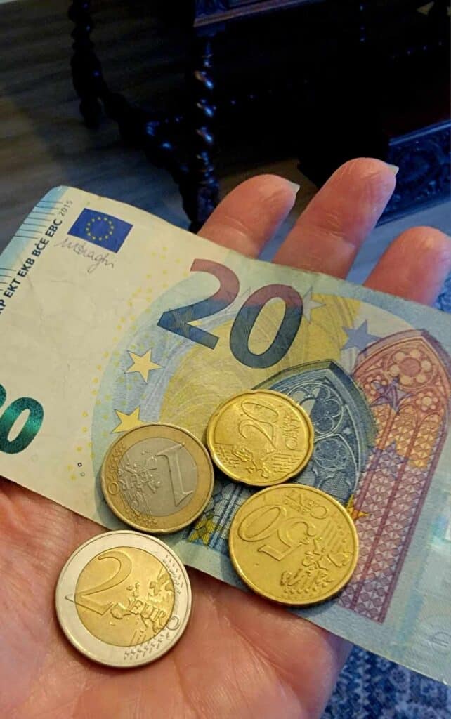 A woman't hand holding a 20 euro note, and four coins - 2 euros, 1 euro, 50 cents, and 20 cents