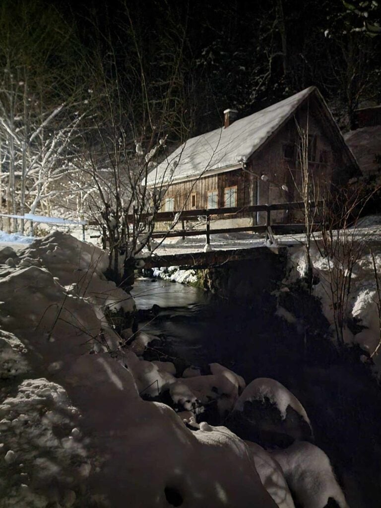 A stone millhouse with windows and snow on the roof. A wooden bridge crosses a stream which flows over large snow-covered boulders.