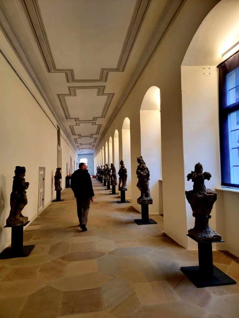 A man walking in a long gallery with statues on either side and windows on the right